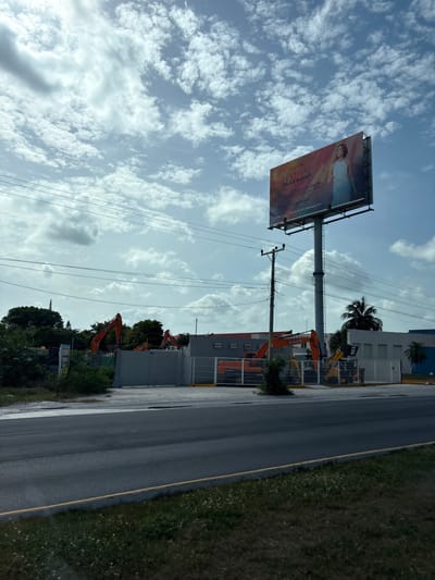 This image depicts a construction site in Cancún, Mexico, as evidenced by the presence of several excavators. A prominent billboard advertising the "V Edición Festival Paax GNP" is visible in the upper right quadrant. The festival is scheduled for July 23rd to August 2nd, 2026. The sky is partly cloudy with a mix of sun and clouds. Power lines and a utility pole are visible in the foreground, suggesting an urban or suburban setting. The construction site is enclosed by a fence, and some landscaping is present around the perimeter. The road in the foreground appears to be a multi-lane highway.