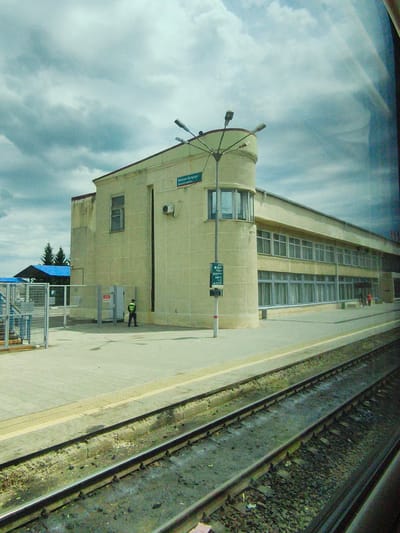 The image shows a train station on a cloudy day. A light-colored, modern-looking building with large windows is the central subject. A rounded tower-like section juts out from the building's corner, topped with lights. Above this section, a sign reads "ВОКЗАЛ БУЗУЛУК" (Buzuluk Station) in Russian, with smaller English text below it.

In front of the building, a paved platform runs alongside train tracks. A person in a high-visibility vest and dark pants stands near a gate, seemingly on duty. A blue-covered canopy and a fenced area are visible to the left of the building. In the distance, several evergreen trees can be seen. The sky is filled with dramatic, dark clouds, suggesting overcast or potentially stormy weather. The time of day is difficult to ascertain due to the cloud cover, but the lighting suggests it is daytime. The scene conveys a sense of calm, perhaps a quiet moment at a regional train station.
