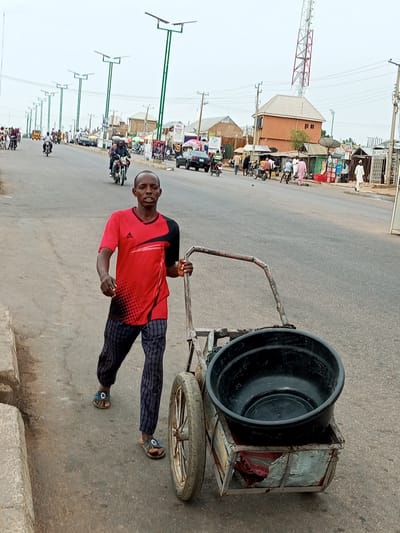 In Tumfure, Nigeria, a man is pushing a cart on a street. He is wearing a red and black t-shirt, striped pants, and sandals. The cart has two wheels and a metal frame, carrying a large black basin. The street is paved, and there are buildings and shops on the right side of the road. Several motorcycles and a few cars are visible on the street, along with other pedestrians. Solar-powered streetlights are also present. The sky is overcast.