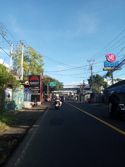 The image captures an urban street scene under a clear blue sky, illuminated by daylight. The asphalt road features a broken yellow centerline, separating traffic lanes, and exhibits minor surface patching.
On the left, a dirt and grassy verge precedes a low concrete wall adorned with graffiti and an advertisement for "Brighton." Behind this wall stands a building with a large red sign for "Prinsesse 2 Hotel & Resort," advertising "Restaurant & Cafe," "Karaoke," "Ball Room," and "Meeting Room." A single dark-colored scooter is parked on the sidewalk near this building. Utility poles carrying multiple power lines line this side of the street, amidst green trees and foliage.
On the right, various structures and shops are visible. A prominent, vertically oriented sign for "MIE GACOAN" with "MIE PEDAS" (Spicy Noodles) is displayed, featuring a pink, blue, and white neon aesthetic. Numerous overhead power lines are densely intertwined across the upper portion of the frame, supported by utility poles. Further down the road, an elevated structure, likely an overpass or bridge, is visible, with additional buildings lining the street beyond it.
Traffic primarily consists of motorcycles, all depicted moving away from the viewer. In the foreground on the right, one motorcycle carries two individuals, both wearing helmets; the passenger is dressed in a dark jacket. Further ahead in the same lane, a second motorcycle is ridden by a single individual wearing a white helmet and light-colored clothing. Additional motorcycles are observed further in the distance, indicating ongoing vehicular movement. The scene is located in the city of Khouathineung, Laos.