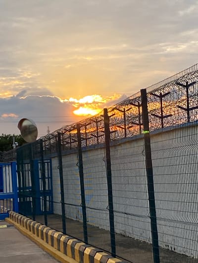 The image depicts an outdoor scene at sunset, ostensibly within the city of Sabon Gari Nassarawa, Nigeria. The sun, a bright golden-orange orb, is partially obscured by clouds on the horizon in the upper-mid to right portion of the frame, casting a warm glow across the sky and illuminating the upper elements of a perimeter fence.
In the foreground, a paved surface occupies the bottom-left, leading to a decorative curb or barrier composed of alternating dark gray/black and yellow/orange hexagonal blocks, which extends diagonally across the bottom of the frame from left to right. To the far left, a portion of a blue metal gate or barrier is visible.
A dark green, possibly coated, mesh fence with vertical support posts runs parallel to the curb, extending into the mid-ground. Behind this fence, a lighter-colored, likely concrete or masonry, wall forms a continuous barrier. Mounted above the wall on the left side is a large, spherical, reflective convex security or traffic mirror. Behind the mirror and wall, a small amount of green foliage is visible.
Topping the wall is a robust security fence system. This system consists of dark, vertical posts with angular supports, holding multiple strands or panels of wire mesh, possibly featuring barbed or razor wire elements. The setting sun directly backlights this security fence, causing its metallic components to glow intensely orange and yellow, creating a silhouette effect against the brighter sky. In the distant background, a faint utility pole or tower is discernible to the left of the sun. No people or active interactions are visible within the frame.