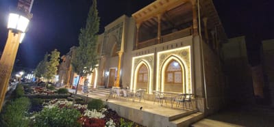 The image captures a nighttime view of a building in the city of Satkak, Uzbekistan, adorned with traditional Uzbek architectural elements. The building features intricate geometric patterns and decorative tilework around arched windows, which are illuminated by warm, yellow light. Above the ground floor, a wooden balcony with carved columns and railings is visible, suggesting a blend of historical and modern design.
In front of the building, there is a paved patio area with several outdoor tables and chairs, set up for dining or relaxation. A well-maintained garden with a variety of flowers in shades of red, white, and purple occupies the foreground, leading the eye towards the building. The garden is illuminated by strategically placed spotlights, enhancing its visual appeal.
On the left side of the image, a tall lamppost with an ornate design stands, contributing to the ambient lighting of the scene. In the background, other buildings and streetlights can be seen, indicating a lively urban environment. A few people are visible in the distance, suggesting the area is a popular spot for evening strolls or dining. The overall atmosphere is inviting and showcases the cultural heritage of the location.