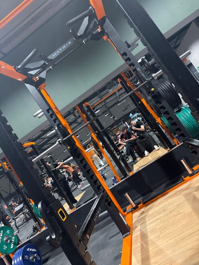 A tilted view showcases a gym interior with several squat racks featuring black and orange frames, along with weight plates. People are visible in the racks and on the floor. The racks are labeled "MATRIX". The floor appears to be a mix of wood and darker material. The image has an unusual angle that creates a sense of depth and complexity.