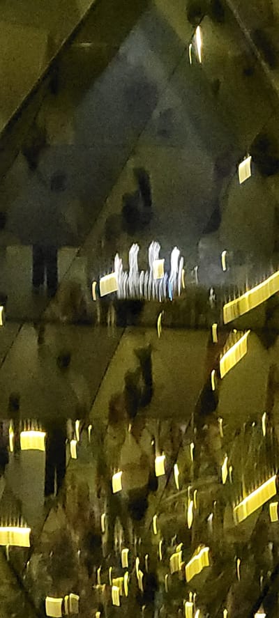 The image depicts an interior space with geometric architectural features, likely a modern building or art installation. The lighting is dim and artificial, with numerous rectangular and elongated yellow lights scattered throughout the scene, creating reflections. In the upper portion of the image, a group of figures, possibly people, are visible as blurred, elongated white shapes with some yellow elements. They appear to be standing in a line or cluster. The overall impression is one of abstract light and shadow, with a sense of depth created by the reflections. The location is identified as Wattens, Austria, which is known for its Swarovski Crystal Worlds, a popular tourist attraction featuring crystal art installations and architectural wonders. Given the abstract and reflective nature of the image, it is plausible that this photograph was taken within the Swarovski Crystal Worlds.
