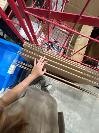 In Holon, Israel, within a storage facility, a person's left hand rests on a stack of flattened cardboard boxes. The boxes are oriented horizontally and are of a uniform size. To the left of the boxes is a bright blue plastic bin. Behind the boxes, a red metal shelving unit is visible, filled with more boxes. The floor is concrete. The person is wearing striped pants and white sneakers. The lighting suggests an indoor environment, possibly a warehouse or industrial space.