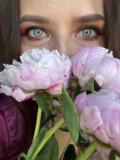 This close-up, vertically oriented image captures a person's face, primarily their eyes and forehead, partially adorned with a bouquet of peonies. The person has fair skin and dark brown hair, which frames the face and is visible on the left and bottom right sides. Both eyes are open, revealing striking blue-green irises. They are accentuated with dramatic makeup, featuring dark eyeliner and reddish-brown eyeshadow, particularly noticeable along the lower lash line. The eyebrows are dark and well-defined.
Dominating the right side of the frame are several large, lush peonies. Their petals display a delicate light pink color that fades into white, with hints of deeper pink along some edges. Green stems and leaves are interspersed among the blossoms, providing a vibrant contrast. A small portion of dark purple or maroon fabric, possibly clothing or a background element, is visible in the lower right corner. The lighting is soft and even, highlighting the intricate textures of the flower petals and the detailed eye makeup. The overall mood is contemplative, with no specific activity beyond posing for a stylized portrait. No text is visible in the image. The photo originates from Rusalevka, Russia.