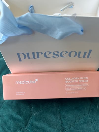 The image shows a box of Medicube Collagen Glow Booster Serum. The box is peach colored and has white text. The text indicates that the serum is "Dedicated to real results" and contains "Hydrolyzed Collagen-Elastin" and "Milk Protein Extract 1%". The box also states it is 15 ml / 0.50 fl. oz. Above the box is a white gift bag with light blue ribbon handles. The background is a dark green textured surface, possibly a blanket or fabric. The scene is indoors and lit by artificial light, creating a shallow depth of field. There are no people or animals visible in the image, nor any indications of its location in Liverpool, United Kingdom.