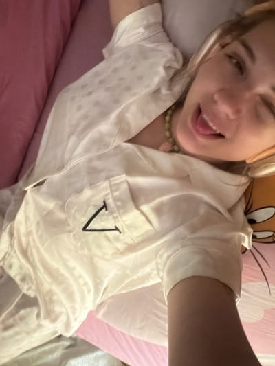 A fair-skinned person with blond hair is lying on a pink bed in what appears to be an indoor location, possibly in Большая Шильна, Russia. The person is wearing a white satin pajama top with a black "V" embroidered on the pocket, and a green beaded necklace. The person is winking and sticking out their tongue. Behind the person is a pillow with a cartoon character on it.
