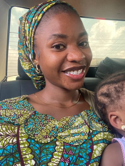 A woman with dark skin and a bright smile poses in what appears to be a car. She is wearing a patterned headscarf and a green and blue patterned top. A gold necklace is visible around her neck. Resting on her shoulder is a baby with curly hair. The baby is wearing a pink ruffled garment. A vehicle window is visible in the background.