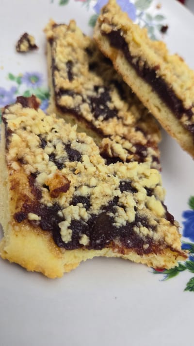 A close-up, eye-level shot shows three pieces of jam-filled crumble bars arranged on a white plate with a floral pattern in blue and green. The bars are layered with a golden-brown crust, a dark red jam filling, and a crumbly topping. One bar is positioned in the foreground on the left, with another slightly behind it to the right. A third bar is angled in the upper right corner, showing a side profile. A small, detached crumb rests in the upper left quadrant of the image. The lighting is bright, highlighting the textures of the baked goods and the plate's design. The location is a domestic setting in Kaka, Turkmenistan, likely a kitchen or dining area.