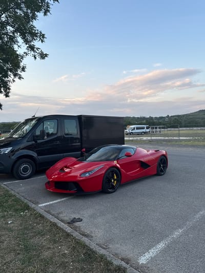An outdoor scene captured in Mogyoród, Hungary, depicts a parking or staging area. A red Ferrari LaFerrari, featuring a black roof, black wheels with yellow brake calipers, is centrally parked on an asphalt surface, oriented towards the left foreground. To its immediate left, a black Mercedes-Benz Sprinter van with a solid black cargo box is partially visible. In the mid-ground, a white passenger van is discernible behind a wire fence and distant vegetated hills. The asphalt surface is bordered by a concrete curb and a strip of dry grass. A single dark-colored glove lies on the asphalt near the curb in the foreground. The lighting suggests late afternoon or early evening under a sky with scattered clouds.