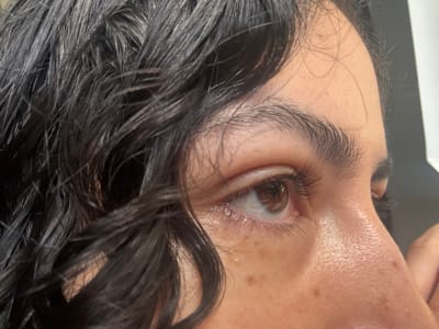 A close-up profile captures a person's face, showcasing a dark, wet, curly hairstyle framing the forehead. Dark eyebrows accentuate the eyes, which are directed to the right. The skin tone is fair, with visible freckles dotting the cheek. The nose is slightly prominent. The lighting illuminates the facial features, creating subtle shadows and highlighting the texture of the hair and skin.