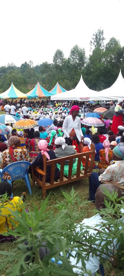 The image shows a large gathering of people in a field in Lubao, Kenya (latitude: 0.379, longitude: 34.78), which is in the Western Region of Kenya.  The individuals are seated on chairs and a wooden bench in front of numerous large tents. There are several people wearing white clothing and sitting in chairs, which are arranged in a line. Some people are standing, including one woman in a white shirt and a pink head scarf, who is standing in the foreground and looking towards the camera. The scene is outdoors, with a green lawn covered in grass. The tents appear to have advertising banners in the background.  The people are seated beneath umbrellas. The scene is likely a gathering for an event or ceremony.