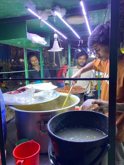 The image depicts a nighttime street food stall in Narayanganj, Bangladesh. The stall is illuminated by multiple bright, linear LED strip lights and a single bare bulb suspended from its green metal frame.

In the right foreground, a man wearing an orange-brown shirt is actively preparing or serving food. His right hand holds a small white bowl with a floral pattern, while his left hand maneuvers a yellow-handled utensil within a large, round metallic pot filled with a yellowish, stew-like substance. A white lid, partially covered by a plastic bag, rests on this pot.

To the immediate right of the large pot, a cooking surface holds numerous pieces of reddish-brown cooked food, possibly fried chicken or a similar meat dish. A small blue gas cylinder is visible beneath this cooking surface.

In the mid-ground, two men observe the activity. One man, positioned left of center, wears a black shirt and looks towards the food. Another man, to his right, wears a light beige shirt and gazes directly forward. In the background, partially obscured, a fourth individual in a red shirt is visible near another section of the stall.

In the lower right foreground, a black bucket contains a murky, greenish liquid with visible particulate matter. A red plastic mug or small bucket is positioned in the lower left foreground. The background is blurred with ambient lights, indicating a bustling urban environment at night.