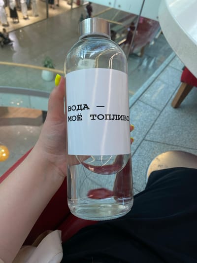 A person's hand with bright yellow nail polish holds a clear glass bottle filled with water. A white label on the bottle displays black text in Russian, reading "ВОДА — МОЁ ТОПЛИВО", which translates to "WATER — MY FUEL". The background is blurred, showing reflections of people and a modern interior, possibly a shopping mall or cafe. The flooring appears to be tiled. The overall lighting suggests an indoor environment. The location is stated to be Kovalevo, Russia.