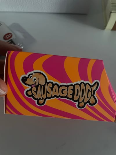 A product box with a "Sausage Dog" logo and a cartoon dog is visible in the foreground. The box has a pink and orange swirl pattern. The background is blurry and appears to be a table or shelf with some shadows. A person's hand is visible on the left side of the image. The scene is indoors. No specific location in Brovary, Ukraine, can be identified.