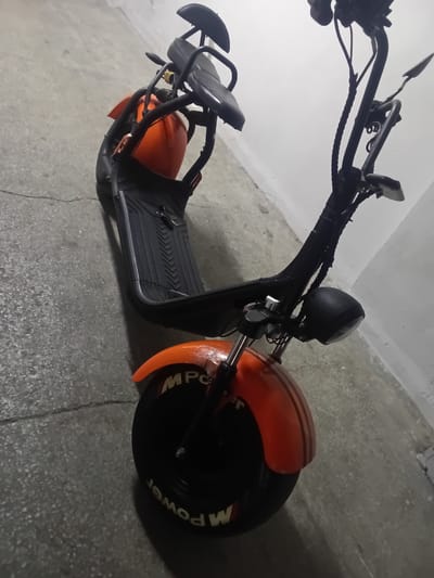 The image shows a black and orange electric scooter parked on a concrete floor. The scooter has a wide frame, a black seat, and a prominent orange fender over the rear wheel. The handlebars are black and include a headlight. Text visible on the rear tire reads "MPower". The setting appears to be an indoor space with a white wall in the background. The lighting suggests it could be indoors with artificial light, or perhaps early morning or late evening. There are no people in the image, and no specific activity is taking place; the scooter is simply parked. The concrete floor shows some cracks and is speckled with small stones, indicating a utilitarian or industrial setting. The overall impression is of a modern, electric scooter in a neutral environment.