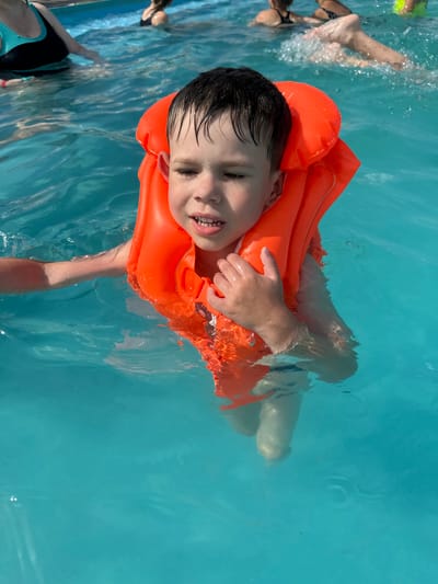 This is a vertically oriented, close-up, overhead shot of a young boy wearing an orange life vest, floating in a bright blue swimming pool. The boy's face is turned towards the camera, and he has dark, wet hair and is smiling with his mouth open, showing his teeth. His right hand is resting on the life vest. His legs and feet are visible submerged in the clear water. In the background, parts of other people are visible in the pool, including legs kicking and a person wearing a black and teal swimsuit. The water is clear and reflects the sunlight, creating ripples and highlights. The scene suggests a sunny day at a swimming pool, likely during a recreational activity or vacation. There is no visible text in the image.