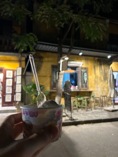 A first-person perspective shot shows a hand holding a small cup of ice cream with a plastic spoon in it. The cup is colorful with cartoonish designs of flowers and animals, and the word "MARIBU" is visible on it. The background features a dimly lit street scene in Hoi An, Vietnam, at night. A weathered, yellow building with a small open window is behind the ice cream cup. The window displays a menu board with an image of ice cream. Several wooden stools are lined up in front of the building, and a closed umbrella stands to the right. The scene is set outdoors on a paved surface, with trees and foliage overhead. The lighting suggests it is evening or night.