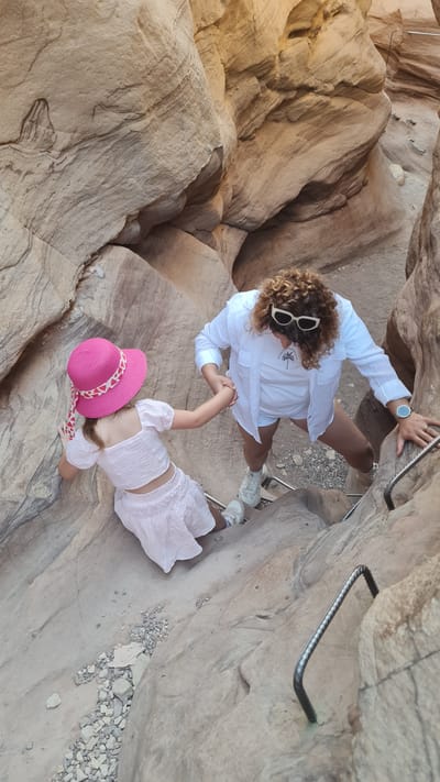 A high-angle, eye-level photograph shows an adult and a child standing on a rocky terrain within a canyon.
The adult, positioned at the top of the frame, is wearing a white button-down shirt, white shorts, and white sneakers. Curly hair is visible, and a watch is on their left wrist. Sunglasses rest on the head. The person is gripping a metal bar installed in the rock face.
The child, wearing a pink hat and a matching pink and white outfit, is holding the adult’s hand. The ground is composed of light-colored, striated rock formations typical of canyon landscapes. Additional metal bars are installed in the rock face, serving as handholds or steps.
The environment suggests a dry, rocky, and possibly desert-like setting. This scene takes place in the Red Canyon, north of Eilat, Israel.