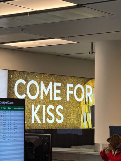 The image shows a large digital display in an airport terminal that reads "COME FOR KISS". The display features a gold textured background and an image that appears to be a detail from Gustav Klimt's painting "The Kiss". To the left of the display is a baggage claim information screen showing flight numbers, belt numbers, and airline information. The screen lists several flights from Austrian Airlines. In the foreground, a woman wearing a red jacket is visible, holding a phone as if taking a photo or video. The environment appears to be an indoor airport luggage claim area, indicated by the presence of the baggage carousel and the information screens. The location is the Vienna International Airport (VIE) in Schwechat, Austria, given the presence of Austrian Airlines flights and the general context of an international airport.