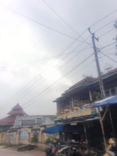 The image displays an outdoor street scene in Dakshin Hichachhara, India, under an overcast sky. A prominent dark utility pole stands on the right side of the frame, supporting numerous overhead power lines that crisscross the upper portion of the image.
To the right, a two-story building features a light yellow/cream colored facade and a red-tiled roof. The second floor includes a balcony with a white railing and visible windows. A sign bearing the word "CENDANA" in black lettering on a white background is mounted above the first floor. Extending from the ground level of this building is a temporary structure with wooden or metal supports, covered by a bright blue tarp. Beneath this tarp, several motorcycles or scooters are parked; a yellow scooter is partially visible on the far right edge of the frame.
Further in the mid-ground, on the left, a building with a distinctive multi-tiered, red-tiled pyramidal roof is visible behind a low wall or fence. Some vegetation is present on the roofs and surrounding the structures. No individuals are discernible within the frame.