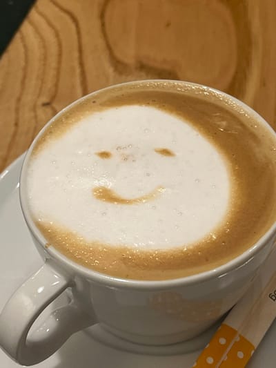 A white ceramic coffee cup, positioned on a matching white saucer, occupies the majority of the frame. The cup contains a beverage with a light brown liquid base, topped by a substantial layer of white foamed milk. A smiley face design is present in the foam, formed by two small, oval-shaped indentations of coffee creating eyes and a curved, thinner line of coffee for a mouth. The cup handle is visible on the left side. To the immediate right of the cup, two elongated paper packets, white with yellow polka dots on their lower sections, are partially visible, consistent with sugar or sweetener packets. The background consists of a light brown wooden surface displaying distinct grain patterns. No individuals are visible. The specific location within the city of Santander, Spain, is not identifiable from the visual content of this image.