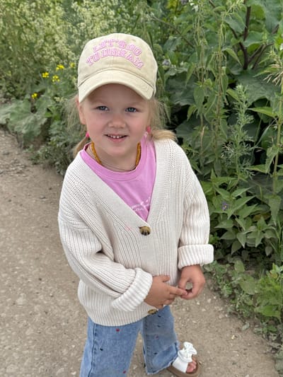A young girl with blonde hair, blue eyes, and a bright smile is pictured outdoors. She is wearing a beige baseball cap with the words "LET'S GO TO DREAM" embroidered in purple letters. She has on a pink t-shirt and a cream-colored knitted cardigan with two brown buttons. Around her neck, she wears a yellow beaded necklace. Her lower half is covered by light blue denim jeans adorned with small red and black embroidered shapes, and a pair of brown sandals with white bows.
The girl is sitting on the ground, which appears to be a dirt path. Behind her and to her left, lush green foliage and plants with small yellow flowers are visible, suggesting a natural, possibly rural or park-like setting. The lighting indicates it is daytime, with soft, diffused light. The overall mood is cheerful and relaxed, with the girl looking directly at the camera with a happy expression.
The provided context specifies the location as "Горный, Russia." There is no specific information about the time of day or weather beyond the visual cues of daylight.