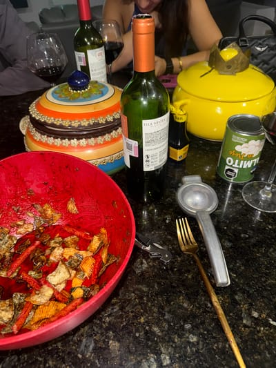 The image depicts a cluttered tabletop scene with various objects. There are two bottles of wine, one with a red label and one with a green label visible. A can of "Palmito en Trozo" sits near a yellow teapot. A silver lemon squeezer and a gold fork are next to a wine opener. A red bowl, filled with chips and what appears to be some kind of sauce, dominates the lower right corner of the frame. A glass of red wine is also visible. Part of a black purse and an arm are on the left side of the image. The background suggests a dark countertop, possibly in a kitchen or dining area. The image doesn't provide information related to the city of Santa Clara Ocoyucan, Mexico.
