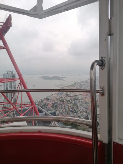View from inside the cabin of a ferris wheel overlooking Hong Gai Ward, Vietnam. The red ferris wheel structure is partially visible to the left, alongside a tall building. The city below is densely populated with buildings, leading to a body of water with islands. The sky is overcast. A metal handrail is present within the cabin. The cabin's interior includes a red seat and a white frame around the window.