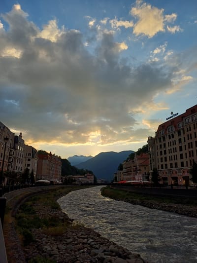This is a landscape photograph taken in Estosadok, Russia, featuring a town nestled beside a fast-flowing river, with a mountain range in the background. The image is oriented vertically.

The scene depicts a developed area with multi-story buildings lining the riverbank. These buildings appear to be hotels or residential complexes, with windows and balconies visible. The river itself is wide and turbulent, with white water indicating a rapid current. The riverbanks are reinforced with stones and greenery.

The sky is a prominent feature, filled with a dramatic mix of clouds. Some clouds are dark and heavy, while others are illuminated by the sun, creating a striking contrast. The sunlight appears to be breaking through the clouds, casting rays and highlighting the edges of the cloud formations. This suggests it is either sunrise or sunset.

The overall mood of the image is serene and majestic, with the natural beauty of the mountains and the river complemented by the human-made structures. The time of day, indicated by the quality of light and the cloud formations, is likely late afternoon or early evening.

There is visible text on one of the buildings that reads "GORKY GOROD" in a stylized font. This is a well-known ski resort and mountain cluster in the Krasnaya Polyana region, near Estosadok.