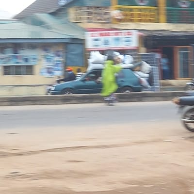 The image shows a street scene in Dadi Riba, Nigeria. A person wearing a bright green outfit is walking along the sidewalk. A turquoise car is parked nearby. Buildings with storefronts are visible in the background, along with signage in local languages. A motorcycle is partially visible at the top of the frame. The road surface is light brown.