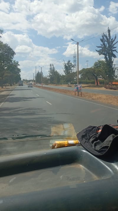 A view from inside a car, showing a straight road stretching into the distance under a partly cloudy sky. Trees line both sides of the road, along with street lights and power lines. Other vehicles can be seen on the road. The car's dashboard is visible in the foreground, holding a rolled cigarette and a dark-colored cap. The dashboard shows signs of wear and dust.
