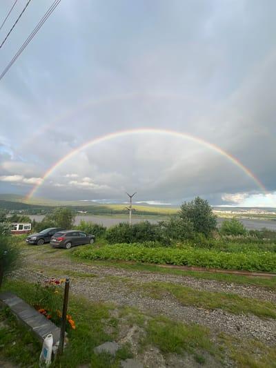 This is a wide-angle, full outdoor shot of a landscape featuring a double rainbow arcing across a cloudy sky. In the foreground, a gravel driveway is visible, leading towards a grassy area with a low stone wall and scattered orange and red flowers. Two cars, a dark gray SUV and a black sedan, are parked on the gravel. Behind the cars, a dense line of green bushes and trees forms a backdrop. Further in the distance, a wide body of water, possibly a river or lake, is present, with a town or city visible on the opposite bank. Hills and mountains form the horizon line. The sky is predominantly gray with some lighter patches of blue and white clouds, suggesting recent or impending rain. The presence of power lines in the upper left corner indicates proximity to infrastructure. The scene appears to be located in or around Zlatoust, Russia, given the description provided.