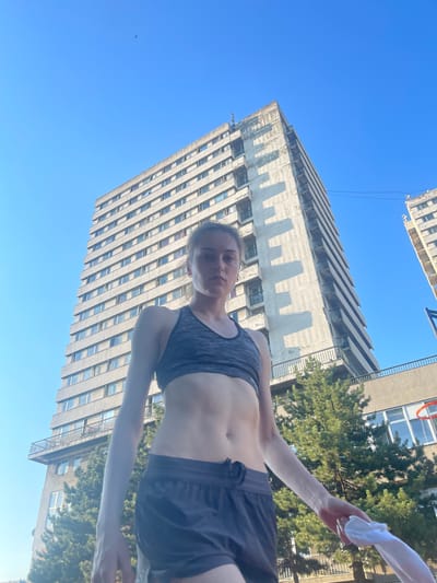 In the city of Palilula Urban Municipality, Serbia, a young woman stands outdoors in front of a tall, concrete apartment building under a clear blue sky. She is wearing a dark grey sports bra and dark grey shorts, and her abdomen is visible. In her right hand, she holds a white cloth. The building, with multiple balconies and windows, shows signs of wear. Green trees are situated in front of the building. To the right, another similar building is partially visible, along with a basketball hoop. The perspective is from a low angle, looking up at the woman and the buildings.