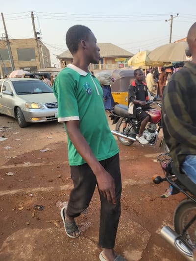 A man in a green shirt and black pants stands on a red dirt road in a bustling outdoor market. Behind him, a silver car is parked. A yellow taxi and several motorcycles are also visible. Other individuals are present, and tents are set up in the background. Overhead electrical wires add to the busy scene.