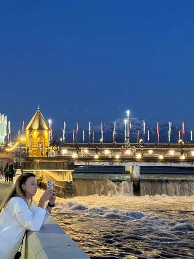 A long shot captured outdoors at dusk shows a woman taking a photo of the Chermenskiy waterfall in Chermen, Russia. A woman in a white shirt is standing on a bridge, holding a smartphone up to take a photo, as a wide waterfall rushes over the structure in the background. Above the waterfall is a walkway with a line of colorful pennants. Beyond that, dark mountains rise in the distance. To the left of the waterfall, a short stone structure with a gold spire and lit windows is also visible. Below the stone structure, a modern white building and other buildings with lights on are present. The sky is a deep blue, suggesting early evening. There are also other people present further away on the walkway and near the woman.