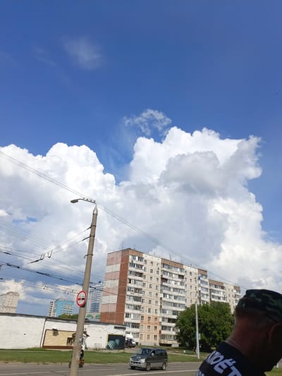 The image captures a street scene in Barnaul, Russia, under a bright blue sky with large, fluffy white clouds. In the foreground, a person wearing a camouflage hat and a black t-shirt with white lettering is partially visible, facing away from the camera. To the left, a concrete lamppost with overhead wires stands prominently. A traffic sign indicating "no overtaking" is affixed to the lamppost. A small electric scooter is parked near the base of the lamppost. A silver minivan is driving on the road, and a grey car is parked on the right side. Behind the lamppost, a long, white building with a yellow section and a mural of a stylized green leaf is situated. Further in the background, multiple multi-story apartment buildings are visible, including a taller, modern blue building and a lighter-colored building with numerous balconies. Trees are scattered throughout the urban landscape. The scene appears to be a typical day in a residential area of the city.