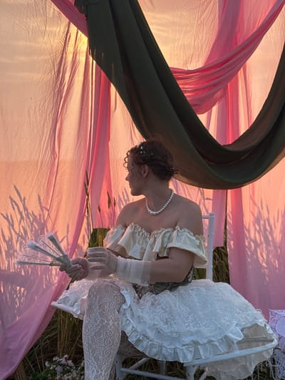 A person, likely female, is depicted in a stylized, almost theatrical pose within an outdoor setting. The subject is wearing a vintage-inspired outfit consisting of a pale yellow, off-the-shoulder top with ruffles, white lace stockings, and sheer white gloves. A pearl necklace adorns their neck. The subject holds a delicate, open fan made of white lace and pale colored metal.

The scene is characterized by flowing fabrics in shades of pink and a dark green. These fabrics are draped and bunched, creating a soft, ethereal backdrop that is illuminated by warm light, suggesting either sunrise or sunset. Shadows of what appear to be wheat stalks are cast across the pink fabric, indicating a rural or field location. Some small wildflowers are visible in the lower right corner. The subject is seated on a white chair, which is partially visible.

The overall mood is romantic and somewhat melancholic, enhanced by the soft lighting and the subject's contemplative posture, with their head bowed. The image conveys a sense of stillness and artistry, possibly a staged portrait or a scene from a narrative. The time of day is likely late afternoon or early morning, given the warm, low-angled light and pronounced shadows. The location context of Foki, Russia, suggests a rural or natural landscape. No text is visible in the image.