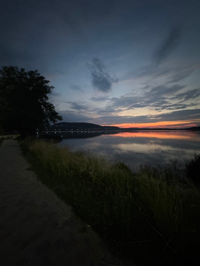 This is a vertical, low-angle photograph of a tranquil sunset scene at dusk. The image is dominated by reflections on a body of water, likely a river or lake, given the presence of reeds and a paved path along the right bank. The water beautifully mirrors the dramatic sky, characterized by streaks of orange and pink from the setting sun, interspersed with dark, moody clouds. The reflection of a distant bridge or structure with lights is visible in the center of the water.

On the right side of the frame, tall grasses and reeds line the water's edge, giving a sense of natural landscape. A paved walkway is visible in the lower right corner, suggesting a park or public area. On the left side, the silhouette of a tree with dark foliage stands against the twilight sky.

The overall mood is peaceful and serene, amplified by the soft light and the stillness of the water. The time of day is clearly dusk, as indicated by the sunset colors and the dimming light. The weather appears calm, with no strong wind suggested by the stable reflections. The location context of Berezhany, Ukraine, suggests a potentially European setting. There is no visible text in the image.
