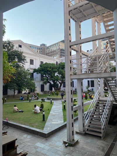 The image captures an outdoor courtyard with a white, multi-story metal staircase structure on the right. Several people are scattered across a manicured green lawn in the center of the courtyard, engaged in activities such as reading and conversing. A white, colonial-style building with dark-trimmed windows is visible behind the lawn. In the background, modern, multi-story buildings rise against a clear sky. The ground is paved with stone tiles. The scene appears to be a public space, possibly a cultural center or a university campus in Kathmandu Metropolitan City, Nepal, given the architectural style and the presence of both traditional and modern buildings.