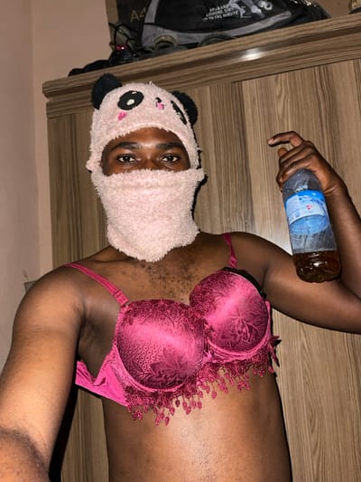 This image depicts an individual in Muchiya, Nigeria, standing indoors, facing the camera directly. The person is wearing a light pink, fuzzy balaclava-style mask or hat with black panda ears and embroidered facial features, covering the head and lower face while leaving only the eyes exposed. A dark pink or maroon bra with textured cups and decorative fringe along the bottom edge is worn on the torso. The individual's right hand is holding a clear plastic bottle that is approximately half-filled with an amber-colored liquid. The background consists of a light-colored wall to the left and a vertical-grain wooden wardrobe or cabinet to the right. Various items, including a dark bag with legible text ("BRAND LEISURE WEAR") and other indistinct objects, are stacked on top of the wooden unit.