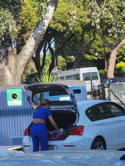 A woman in a blue athletic outfit with her hair tied back in a ponytail is standing at the open trunk of a white sedan, retrieving items from a colorful bag. She has a tattoo on her lower back. The car is parked next to a large blue recycling bin with a green circular aperture for glass disposal. In the background, a white van is visible, along with other vehicles behind a chain-link fence. Lush green trees and foliage surround the scene, suggesting an outdoor setting. The license plate of the white sedan is partially obscured, showing the characters "J28". The environment appears to be a public area in Quarteira, Portugal, likely a recycling point.