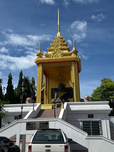 The image shows a gilded Buddhist temple structure in Baan Mae Nam, Thailand. The temple is a golden, tiered building with ornate carvings and a spire at the top. In the central recess of the temple, a large, dark statue of the Buddha is seated. The statue is draped with a yellow sash. The temple is elevated on a white, multi-tiered platform with stairs leading up to it. At the sides of the platform, at the top of the stairs, are two lion statues. The background features a clear blue sky with scattered white clouds, and lush green trees. In the foreground, the rear of a white pickup truck is visible, parked in front of the temple complex. The truck has "Ford" branding on its tailgate. The scene appears to be a bright, sunny day.