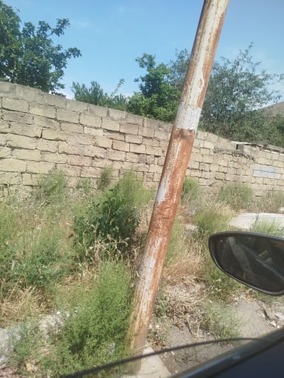 A rusty metal pole stands prominently in the foreground of this outdoor scene. It is positioned to the left of center and is angled slightly. The pole exhibits signs of wear and tear, with patches of white paint peeling away to reveal the rusted metal underneath. Behind the pole, a stone wall constructed from roughly hewn blocks stretches horizontally across the mid-ground. The wall appears aged and weathered.
Beyond the wall, lush green trees and foliage thrive, suggesting a natural or semi-natural environment. The sky above is a clear, vibrant blue with a few faint wispy clouds, indicating a pleasant day. The overall setting appears to be a somewhat undeveloped or rural area, possibly on the outskirts of a city. The presence of what looks like a car's side mirror in the lower right corner suggests that the photograph was taken from inside a vehicle.
The lighting indicates it is daytime, likely late morning or early afternoon, with the sun casting clear shadows. There are no people visible in the image, and no discernable text is present. The scene conveys a sense of everyday life in a place where nature is beginning to reclaim areas around human structures. The arid-looking vegetation in the foreground, contrasting with the greener trees behind the wall, might hint at a drier climate.