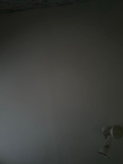 A dimly lit, gray wall dominates the frame. To the left, a patterned, possibly textured material hangs vertically. A two-headed adjustable light fixture is affixed to the upper right portion of the wall. The light fixture and the textures are slightly out of focus.
