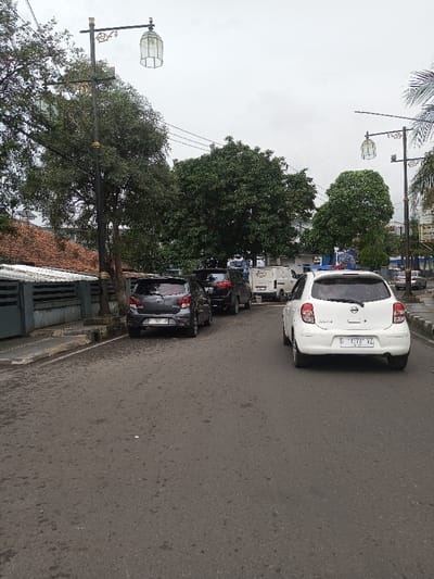 The image depicts an urban street scene under an overcast sky. An asphalt road, appearing damp or stained, occupies the foreground and midground. In the midground, a white Nissan March hatchback is positioned on the right side of the road, facing away from the viewer. Its rear license plate reads "D 1573 VZ". A blue light reflector or accessory is affixed to its rear roofline. On the left side of the road, a dark gray hatchback, possibly a Toyota Agya, is parked parallel to a concrete curb, displaying the rear license plate "F 1321 JN". Behind it, a black SUV is partially visible, also parked. Further into the background, a white utility van and additional vehicles are discernible.
The left side of the street features a low, dark gray concrete wall, behind which a building with a reddish-brown tiled roof and white corrugated metal awnings is visible. Several dense green trees line the street on both sides. Two black streetlights with ornate gold accents and white, hexagonal, cage-like lantern fixtures are prominent; one is on the left side of the road, and another is further down the street on the right. Overhead power lines traverse the upper portion of the scene. No individuals are visible. While the prompt specifies the location as Kodarapalli, India, the visible license plate formats are consistent with vehicle registrations from Indonesia, not India.