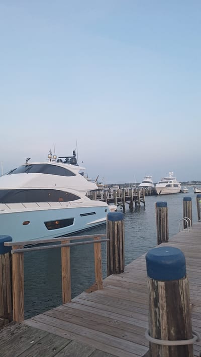 The image shows a view of boats docked at a harbor in Nantucket, USA. In the foreground, a wooden pier with vertical posts topped with blue caps leads into the water. A light blue and white yacht is prominently docked on the left side of the frame. In the background, several other yachts are docked along a pier. The sky is clear and pale blue, indicating it might be early morning or late evening. The water is calm and reflects the light from the boats and the sky. The overall scene depicts a serene harbor setting with luxury yachts.