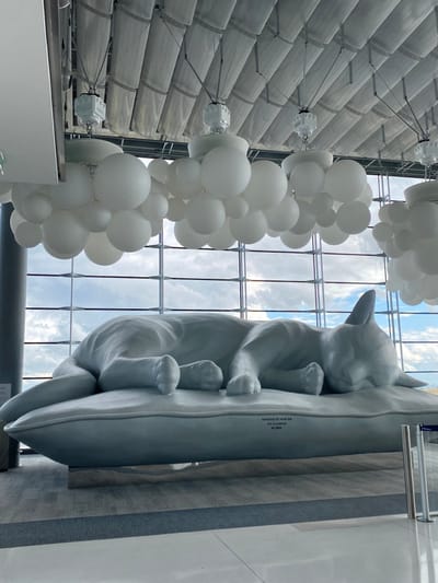 This image captures a large, light-gray sculpture of a sleeping cat on a matching pillow, situated within Terminal 2E of Charles de Gaulle Airport, near Mitry-Mory, France. The oversized cat sculpture is depicted in a relaxed, curled pose with its eyes closed. Text inscribed on the front of the pillow reads "DÉFENSE DE MONTER," "NO CLIMBING," and Japanese characters. The sculpture rests on a low, rectangular base of the same light gray color.
The environment features a high, vaulted ceiling with a white, ribbed or paneled surface. Suspended from the ceiling are numerous clusters of white, spherical light fixtures varying in size, resembling abstract clouds. A prominent multi-paneled glass window wall forms the background, revealing a bright, overcast sky with visible clouds. The floor appears to be a large-tiled or carpeted surface, primarily gray with lighter gray accents. A dark structural column is partially visible on the far left, and a silver stanchion barrier is present on the far right, indicating a public interior space. No people are visible.