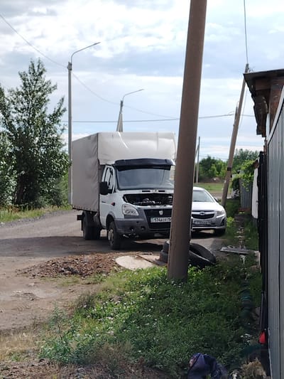 The image depicts an outdoor scene in Ekibastuz, Kazakhstan, under a cloudy sky. In the foreground, a white delivery truck with its hood open is parked on a dirt road. To the right of the truck, a silver sedan is parked. A thick wooden utility pole stands prominently in the center of the image, obscuring parts of the vehicles and the background. To the right of the utility pole, a portion of a building with a corrugated metal wall is visible. The surrounding environment includes green vegetation, a large tree on the left, and scattered debris.
